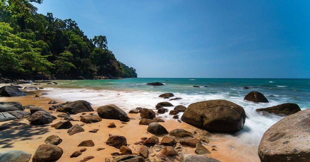 Rocky Khao Lak beach with turquoise Andaman Sea waters, golden sand, and a lush tropical forest along the coastline.